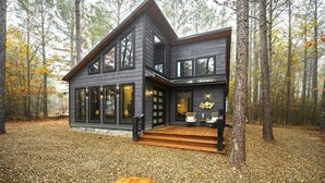 A modern two-story cabin with large windows, a wooden deck, and a forest backdrop. - Moroccan Nights|Hot Tub|Fire Pit|Hammocks (Broken Bow)