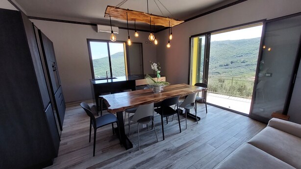 Un comedor moderno con una mesa de madera, sillas e iluminación colgante. Se observa un paisaje montañoso a través de amplios ventanales.