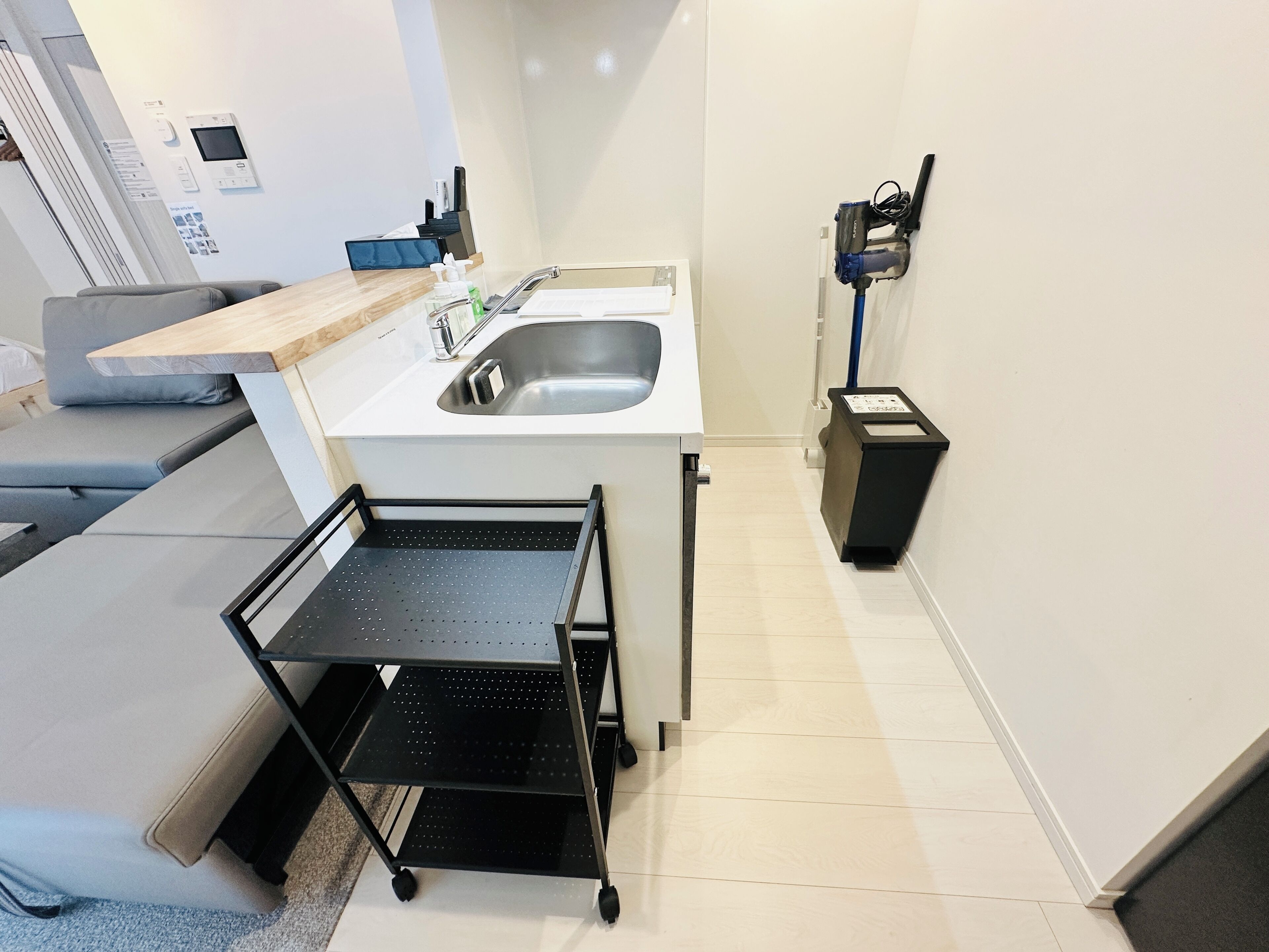 A compact kitchenette with a sink, a small countertop, a black metal cart, and a vacuum cleaner against the wall.