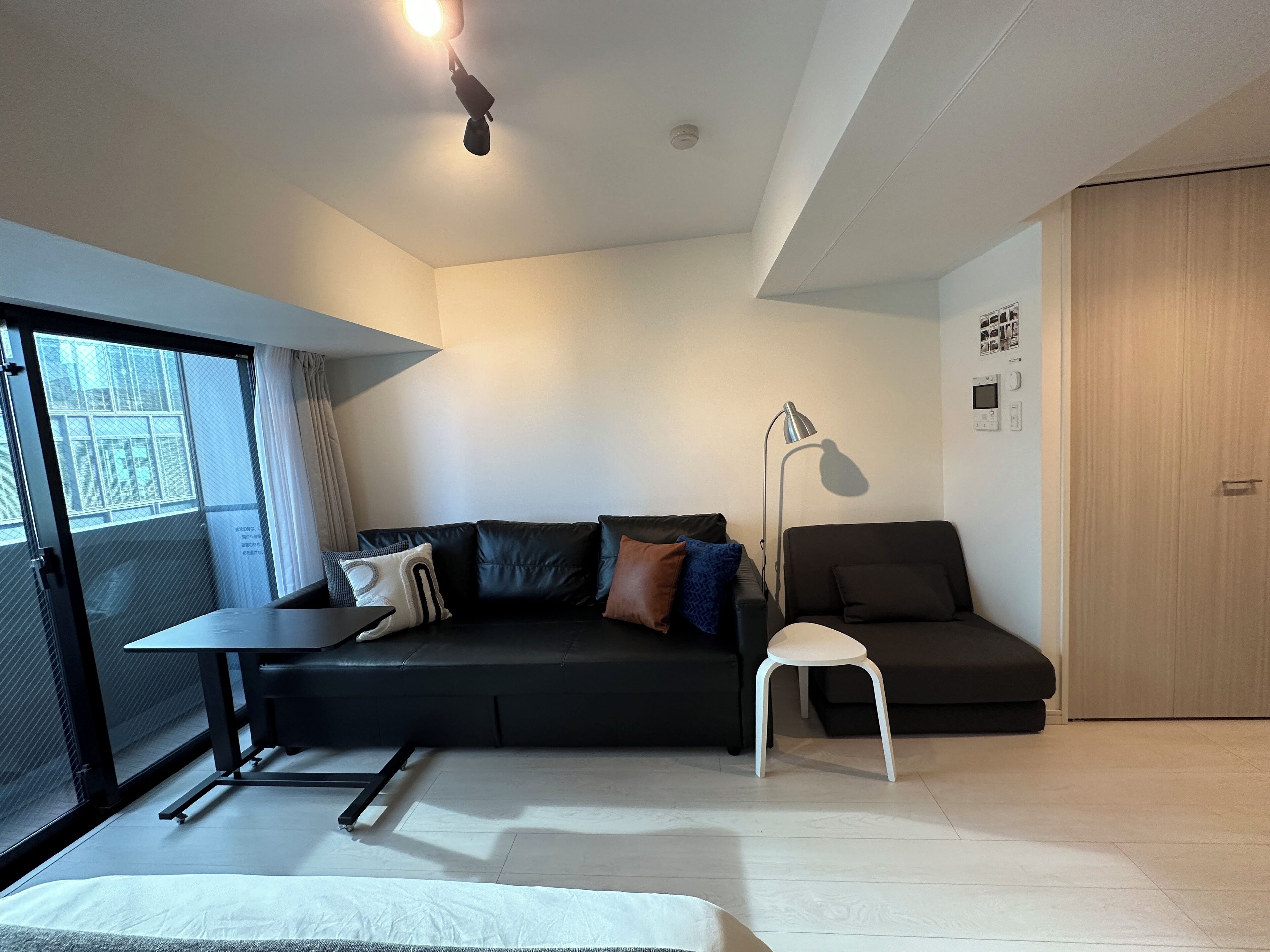 A modern living room with a black sofa, a white chair, a standing lamp, and a large window with curtains.
