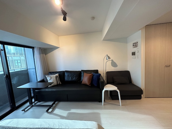 Una sala de estar moderna con un sofá negro, una silla blanca, una lámpara de pie y un ventanal con cortinas.