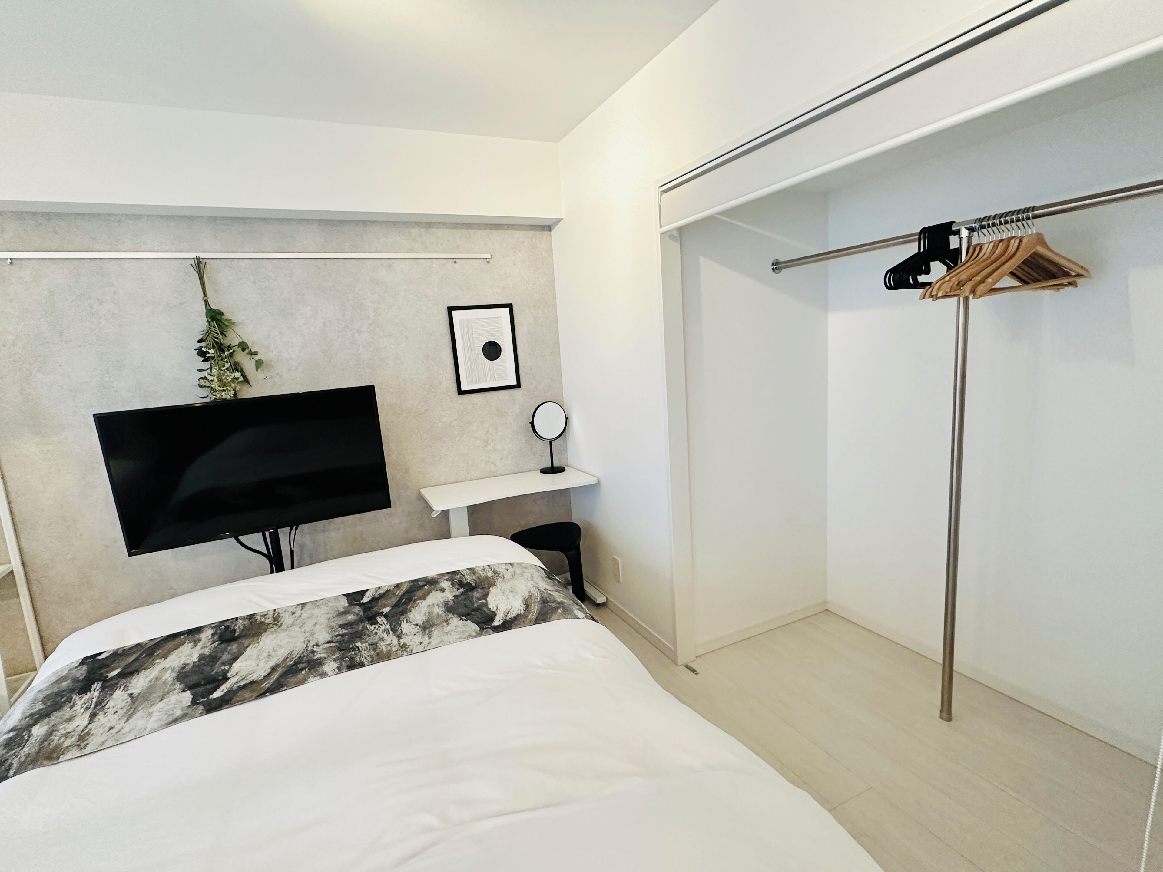 A modern bedroom with a bed, a mounted TV, a small bedside table with a mirror, and a clothing rack.
