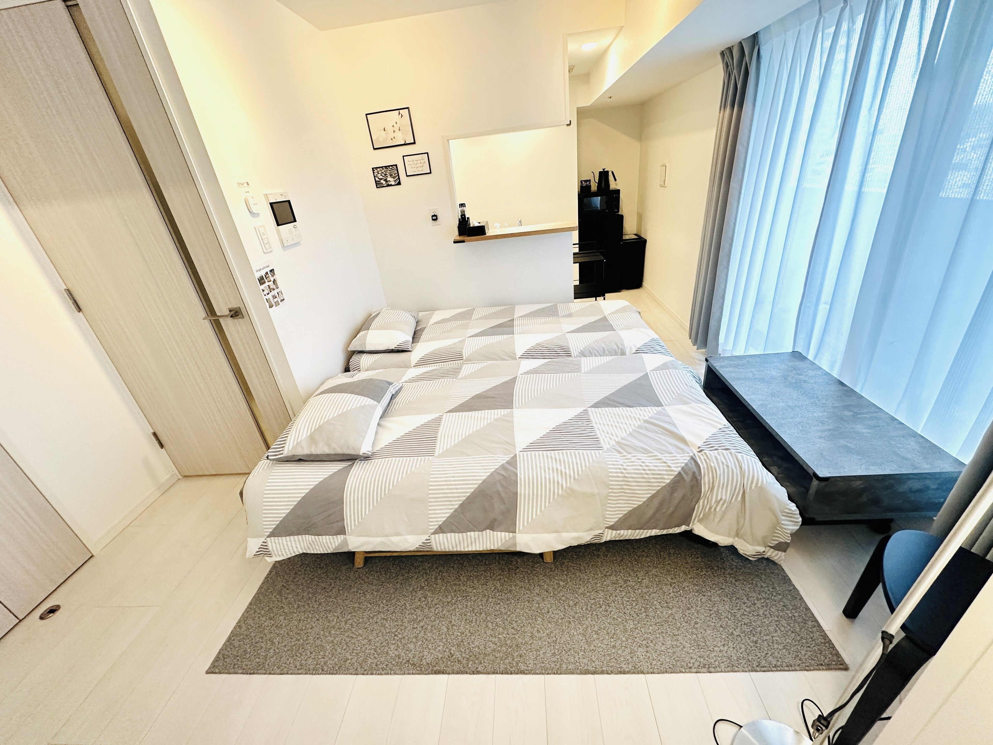 A modern bedroom with a bed, a small desk, a chair, a window with curtains, and a door.