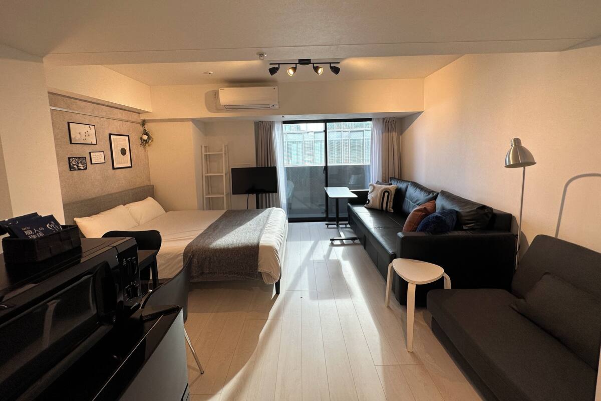 Une chambre d’hôtel avec un lit, un canapé, un bureau et une télévision. Il y a une fenêtre avec des rideaux et une vue sur les bâtiments environnants.