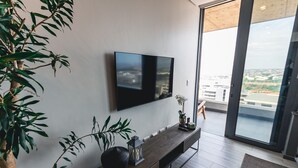 A modern living room with a large flat-screen TV mounted on the wall, a wooden console table with decorative items, and a large window offering a city view. - Umhlanga Arch Luxury Holiday or Work with Sea View (Durban)