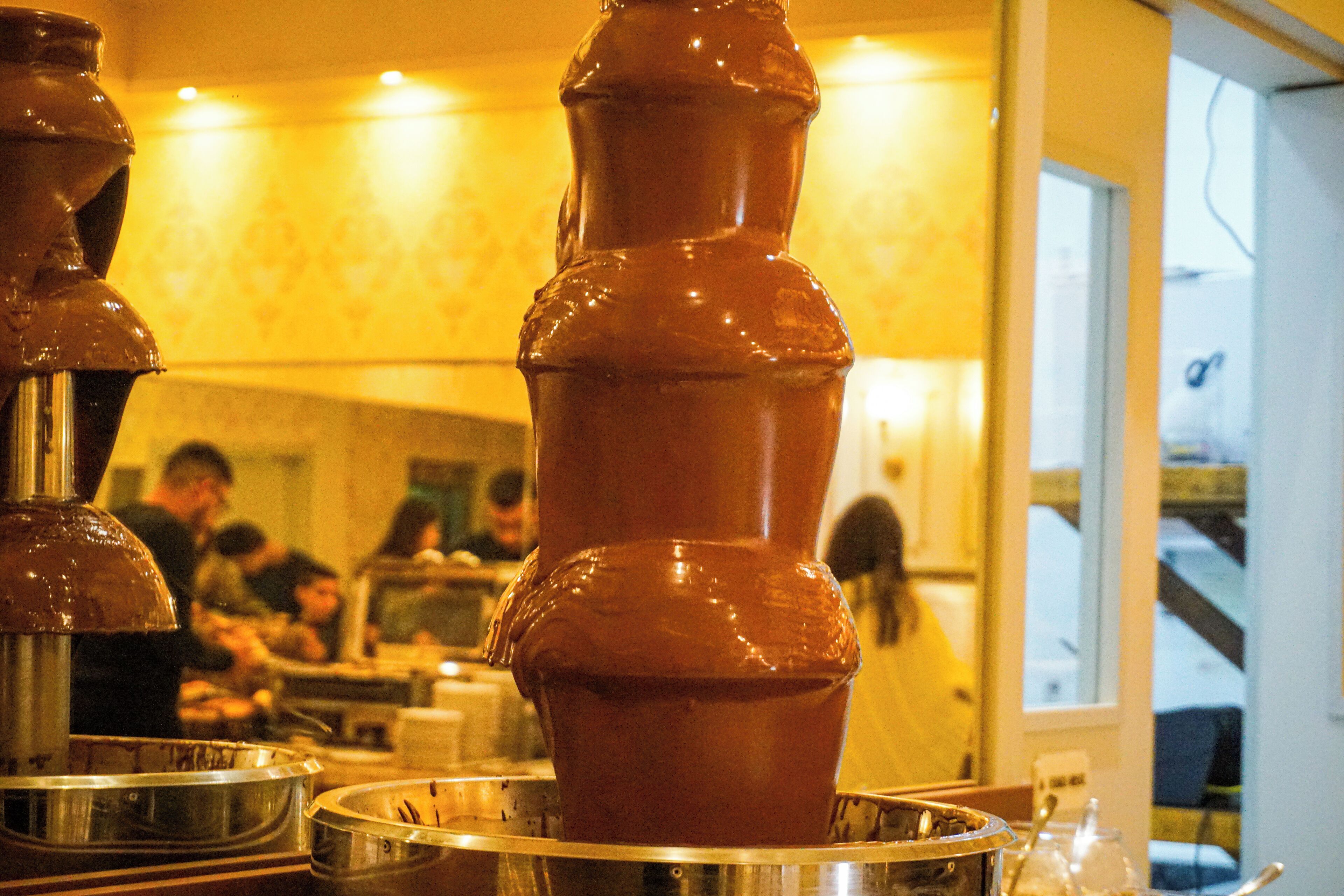 Una fuente de chocolate con varios niveles, rodeada de cuencos metálicos y un área de comedor con gente al fondo.