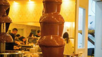 Una fuente de chocolate con varios niveles, rodeada de cuencos metálicos y un área de comedor con gente al fondo.