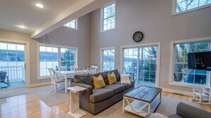 A spacious living room with a gray sofa, a coffee table, and a dining area with a white table and chairs. Large windows provide views of the outdoors.