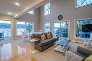 A spacious living room with a gray sofa, a coffee table, and a dining area with a white table and chairs. Large windows provide views of the outdoors.