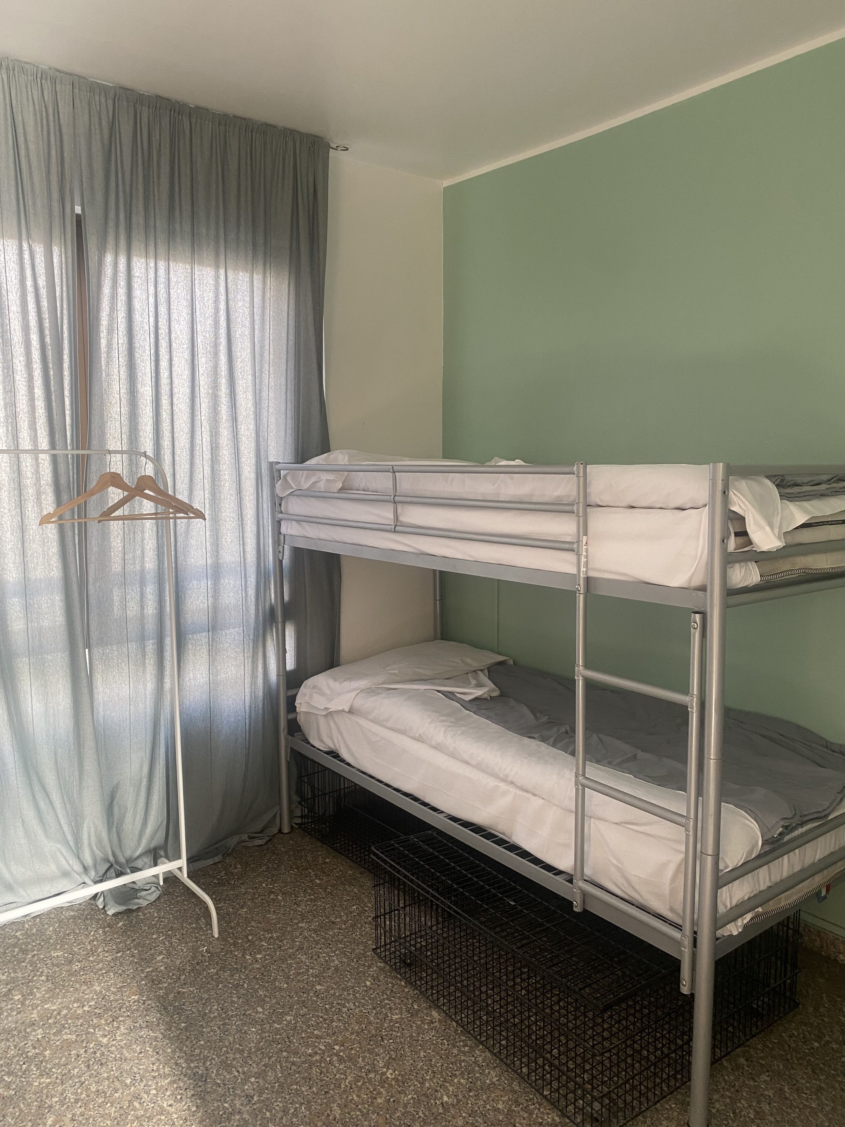 Une chambre avec un lit superposé doté d’une structure en métal, d’un porte-vêtements en bois et d’un rideau.
