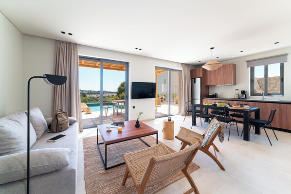 A modern living room with a sofa, coffee table, and wicker chairs. A kitchen area with a dining table and chairs is visible.
