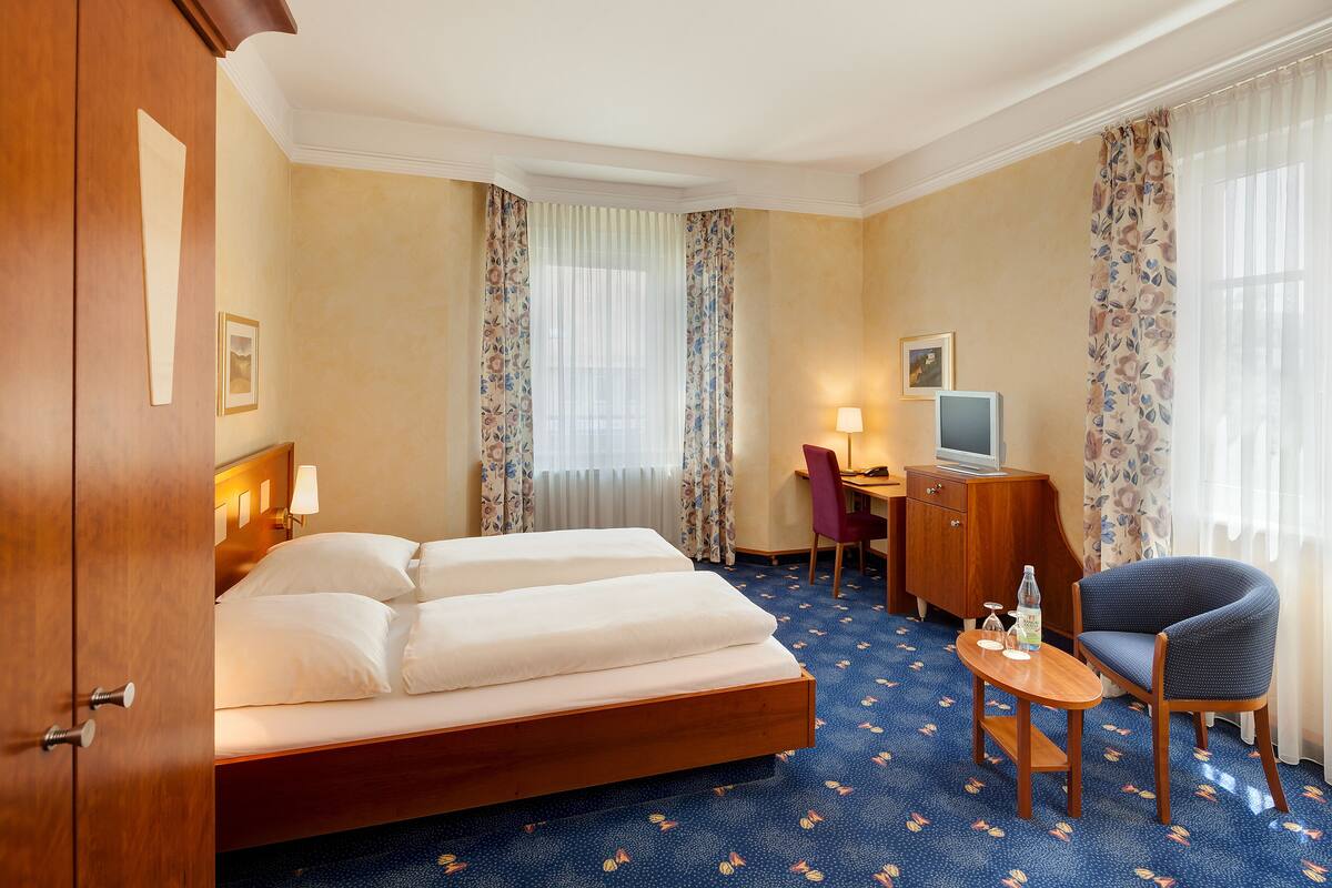 Ein Hotelzimmer mit einem Doppelbett, einem Schreibtisch mit Stuhl, einem Fernseher und einem kleinen Tisch mit Flasche und Gläsern.