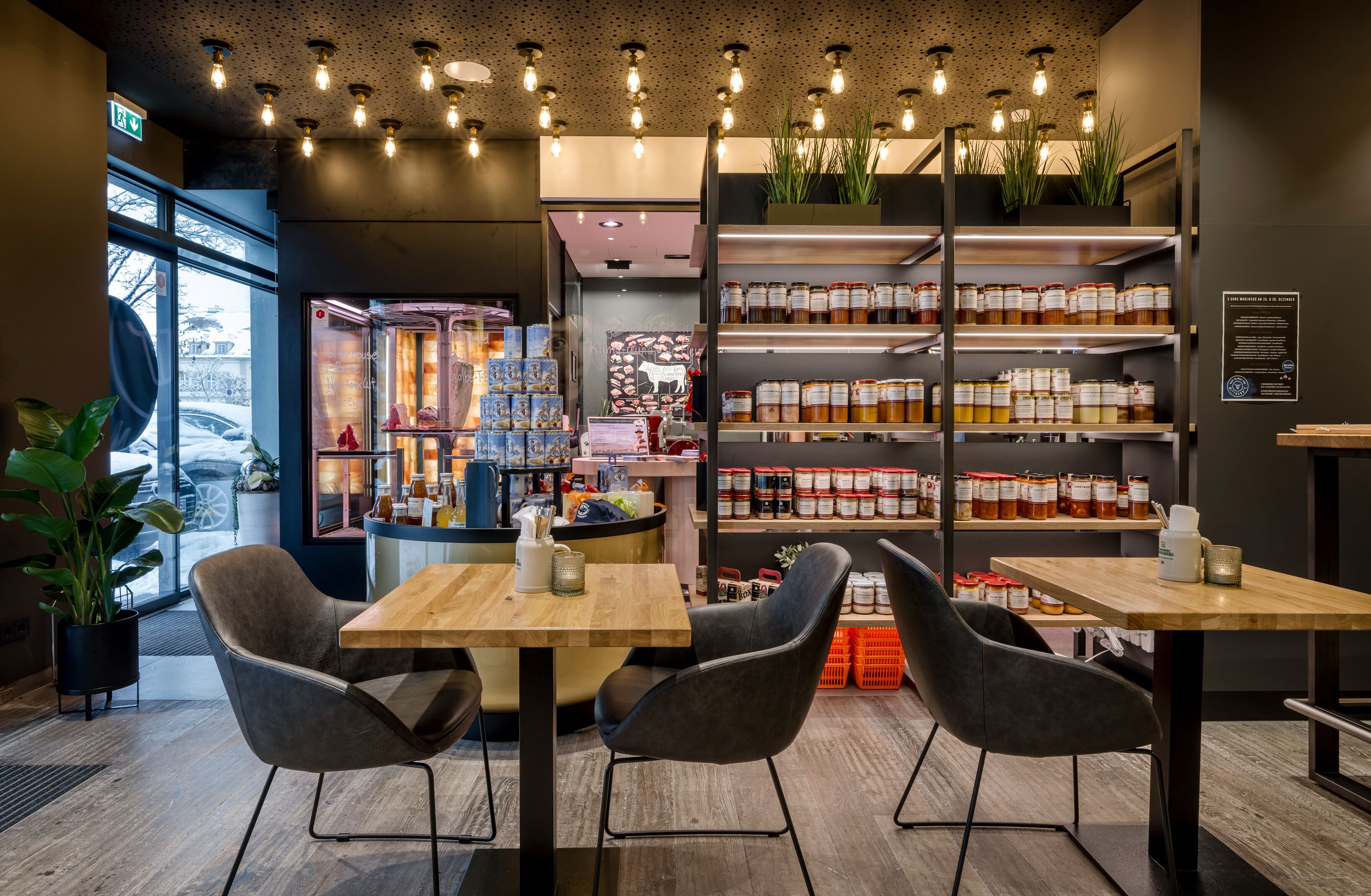 Un interior de cafetería moderno con mesas y sillas de madera, una barra con diferentes productos alimenticios y estantes llenos de frascos con salsas.