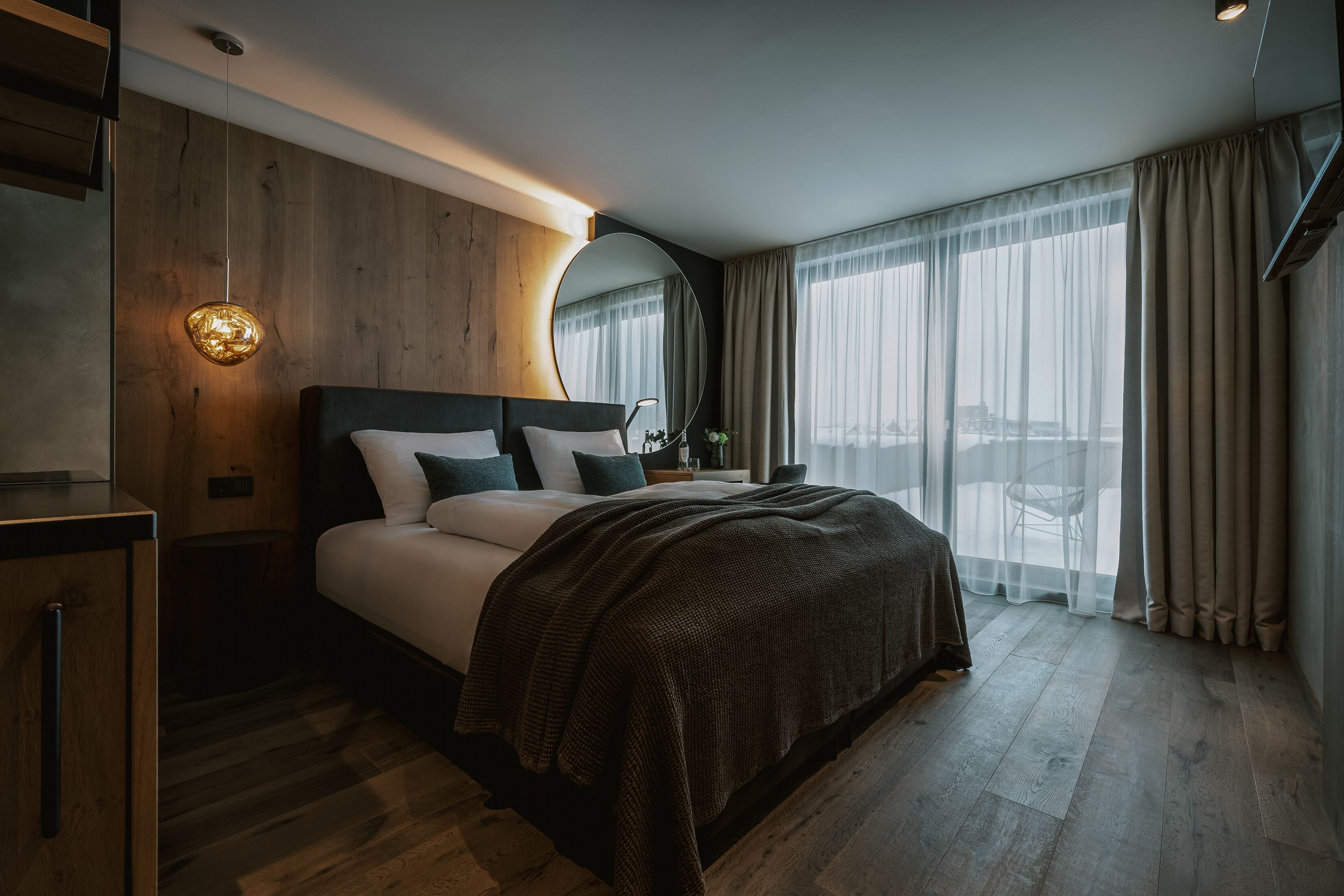 Quarto de hotel moderno com cabeceira de madeira, uma cama grande com manta escura, mesa de cabeceira com luminária, um espelho grande e vista para a cidade através de cortinas leves.