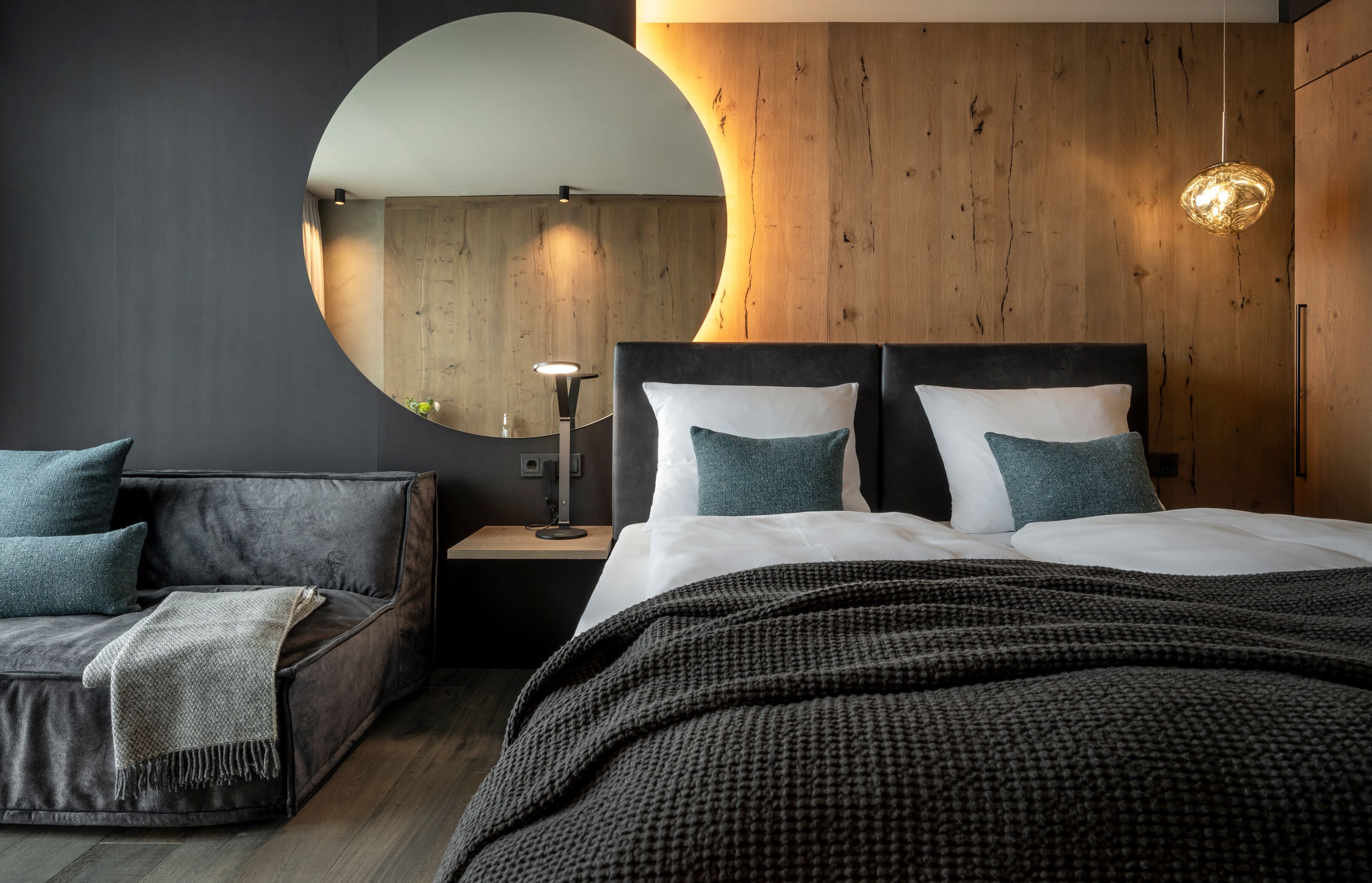 A modern bedroom with a large bed, a round mirror, a wooden accent wall, and a side table with a lamp.