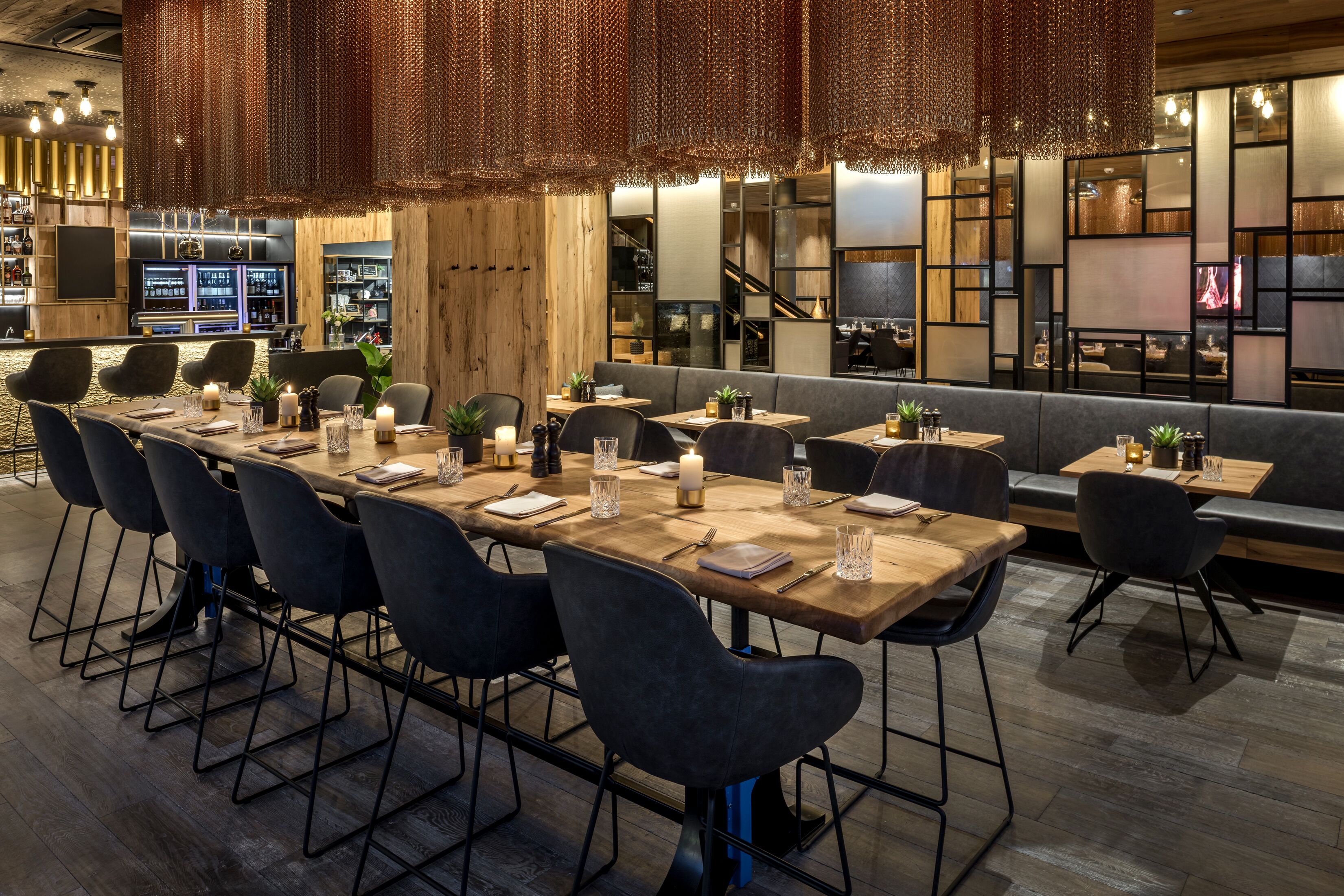 A modern restaurant with long wooden tables and black chairs, a bar area in the background, and a cozy seating area with cushions.