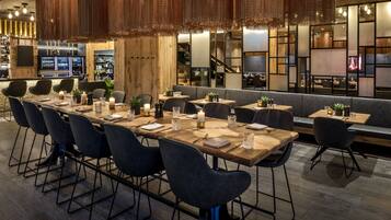 A modern restaurant with long wooden tables and black chairs, a bar area in the background, and a cozy seating area with cushions.