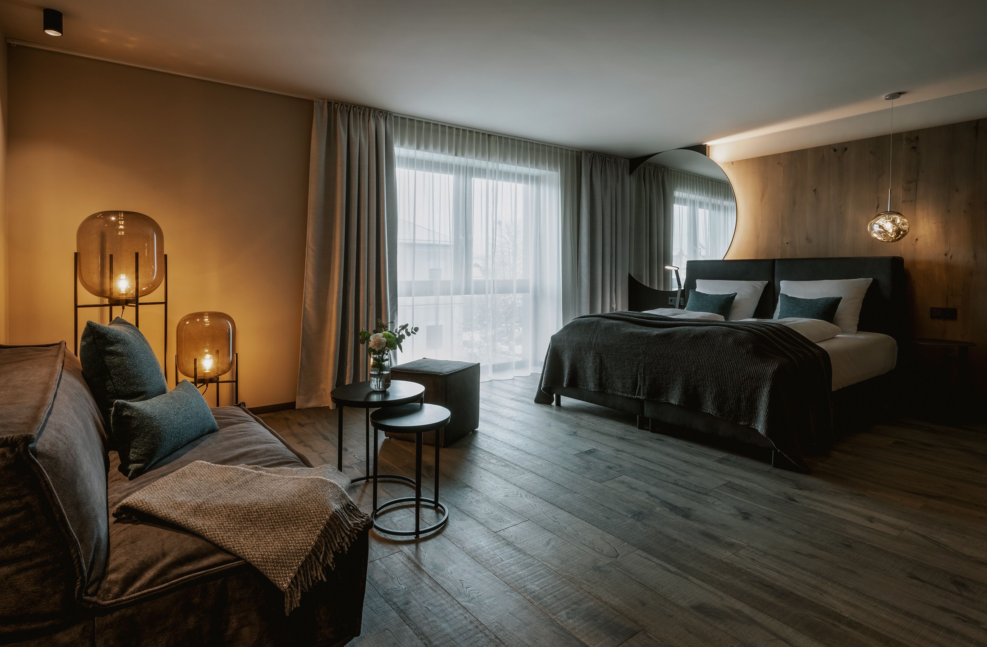 A modern hotel room with a wooden floor, a large bed with dark bedding, a sofa, a small round table with a lamp, and a large window with curtains.