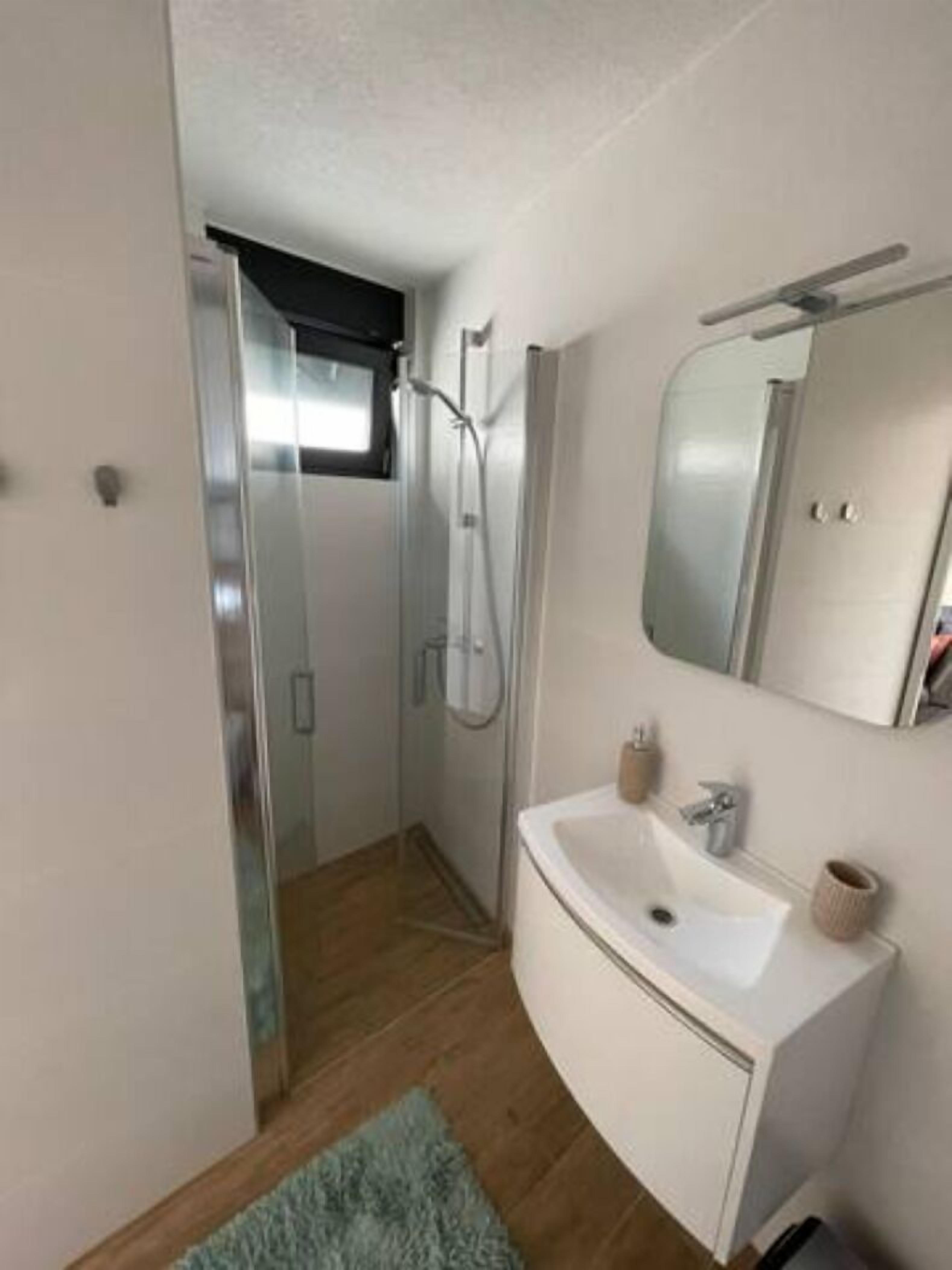 A modern bathroom with a glass shower enclosure, a white sink with a mirror, and a small rug on the floor.