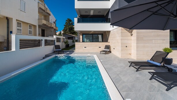 A modern outdoor pool area with a clear blue pool, lounge chairs, and umbrellas. - Villa Laverna Annex (Novalja)
