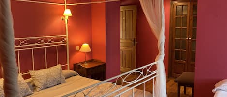 A four-poster bed with a canopy, a bedside table with a lamp, and a wooden door in a room with red walls.