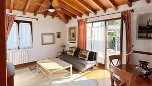 A cozy living room with a wooden ceiling, a sofa with cushions, a coffee table, a dining table with chairs, and a view of the outdoors through large windows. - Posada La Casona de Los Güelitos (Santillana del Mar)