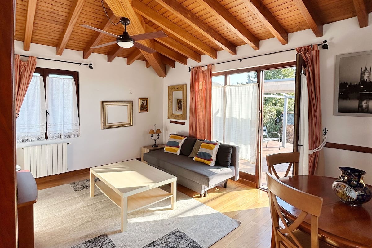 Un salon chaleureux avec un plafond en bois, un canapé agrémenté de coussins, une table basse, une table à manger avec des chaises, et une vue sur l’extérieur grâce à de grandes fenêtres.