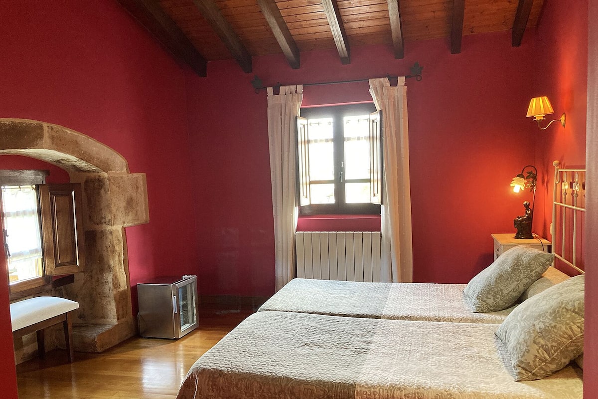 Une chambre avec une arche en pierre, un plafond en bois, un lit avec une literie à motifs, une table de chevet avec une lampe et une fenêtre avec des rideaux.
