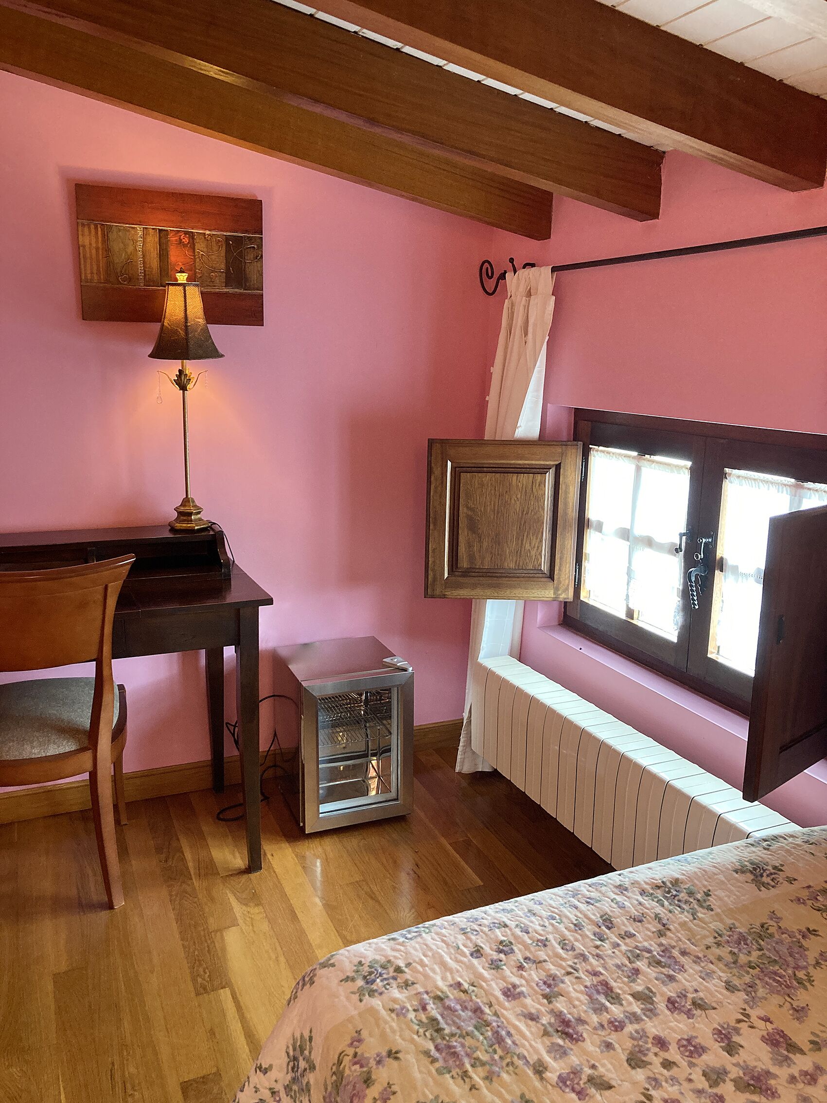 A room with a wooden desk, a radiator, a window with curtains, and a bed with a floral bedspread.
