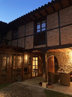 A traditional half-timbered building with a stone archway entrance, wooden shutters, and a stone pathway leading to a seating area with potted plants.