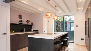 A modern kitchen with a central island, bar stools, pendant lighting, and a view of greenery outside. - Stunning townhouse with large roof terrace (London)