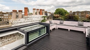 A rooftop terrace with a glass enclosure, a white bench, and a view of surrounding buildings. - Stunning townhouse with large roof terrace (London)