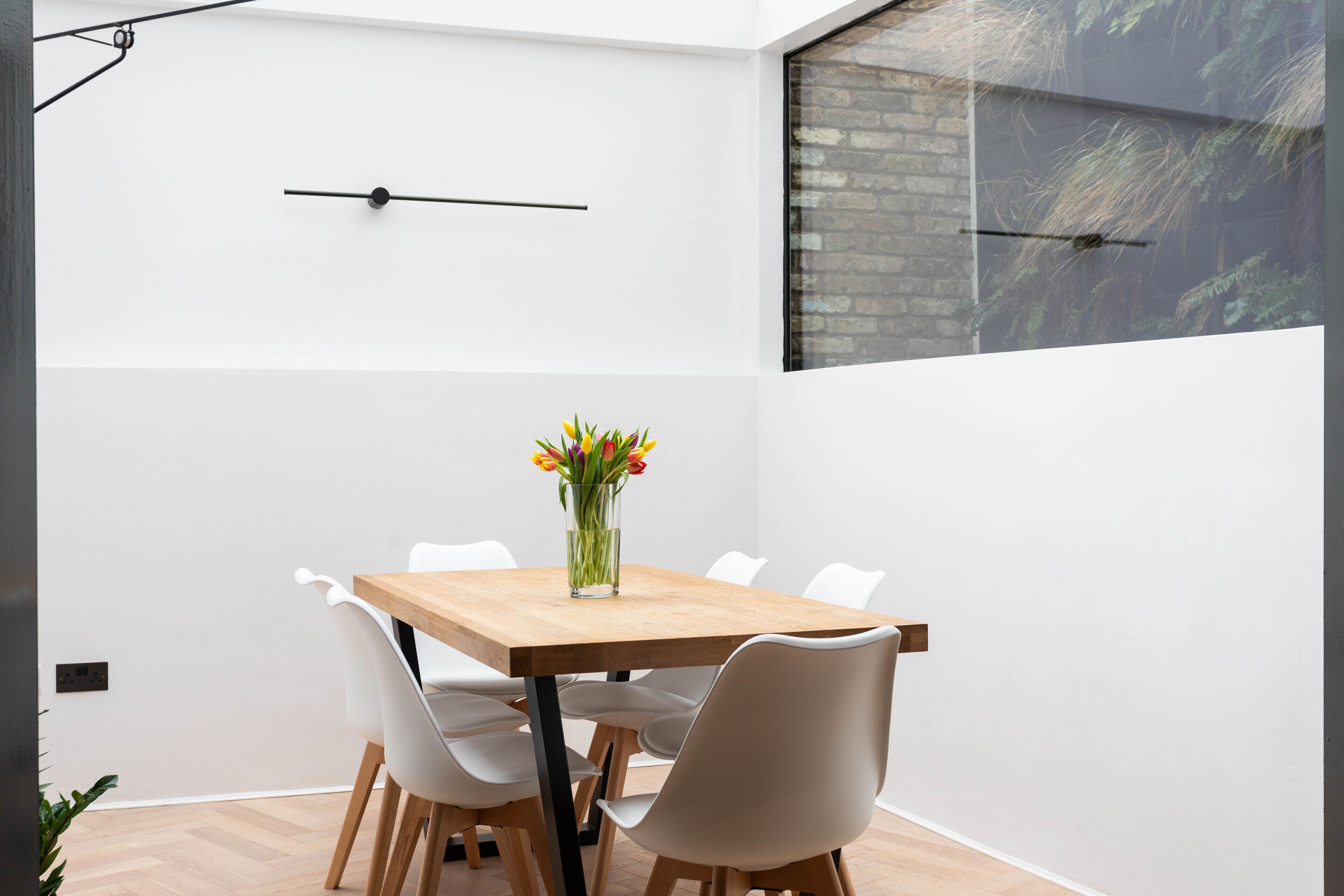 Un espace repas moderne avec une table en bois et des chaises blanches, un vase rempli de fleurs et une horloge noire accrochée au mur.