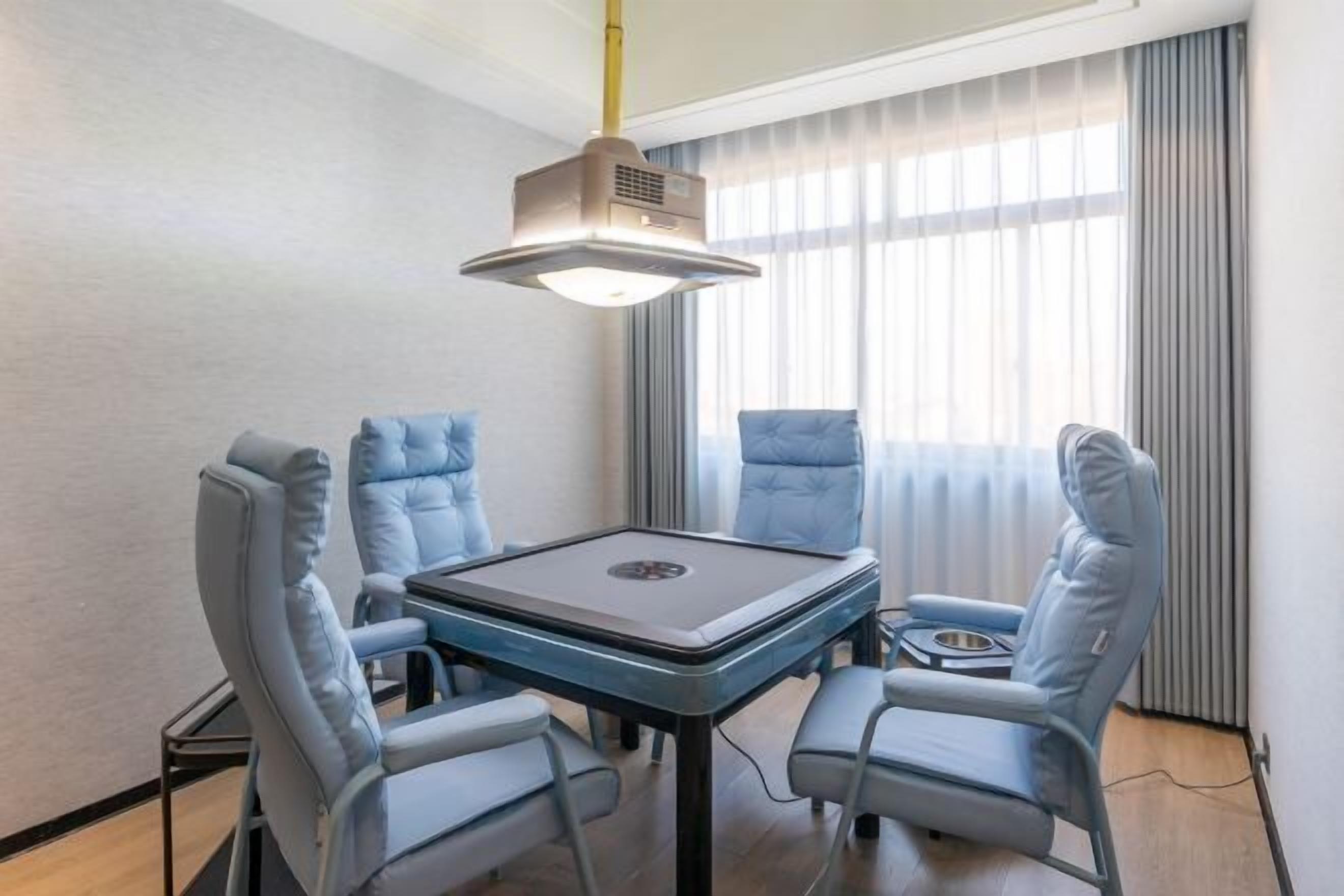Una habitación moderna con una mesa central y cuatro sillas azules, un proyector en el techo y un ventanal con cortinas.