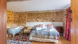 Quarto com parede de pedra, cama com roupa de cama estampada, banheira estilo vintage e chão de madeira.