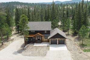 A two-story log cabin with a garage and a covered porch, surrounded by dense forest and a dirt driveway.