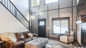 A spacious living room with a brown leather sofa, a wooden coffee table, and a beige armchair. A black door and a large window are visible in the background. - Secluded Cabin on 3 Acres w/ Hot Tub, Open Floor Plan, Starlink Internet! (Lead)