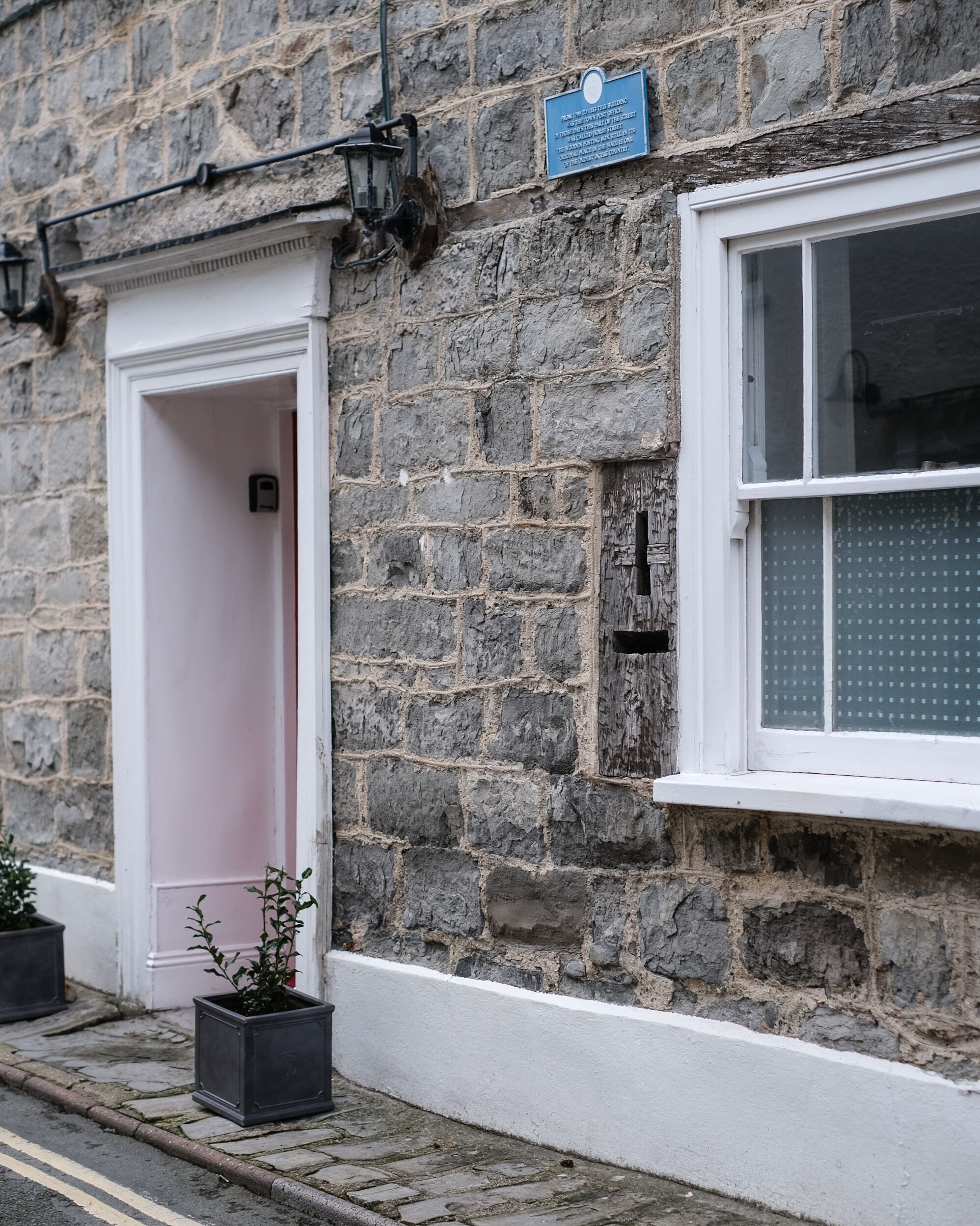 A stone building with a white door and window, a blue plaque, and potted plants on the sidewalk.