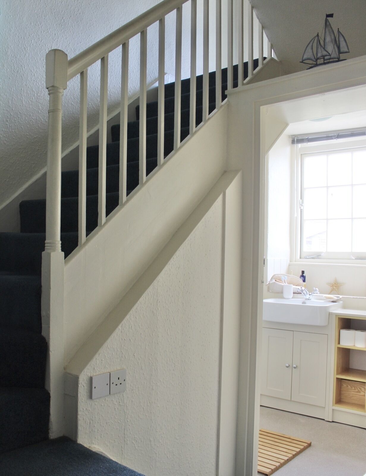 Eine Treppe mit weißen Geländern führt zu einer Zwischenebene, ein Fenster bietet einen Ausblick, und es gibt eine Küchenzeile mit weißen Schränken und einem Holzboden.