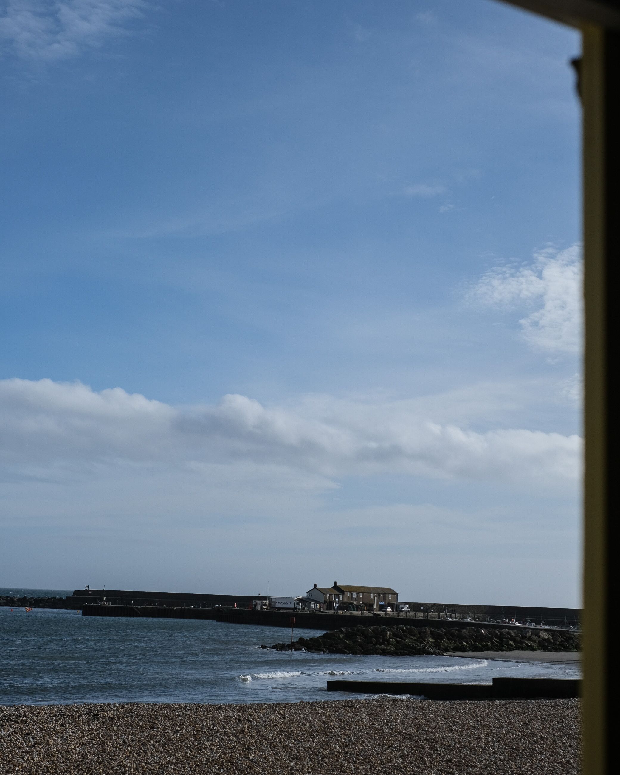 Une vue sur la côte, avec une plage de galets, une jetée et des bâtiments visibles au loin.