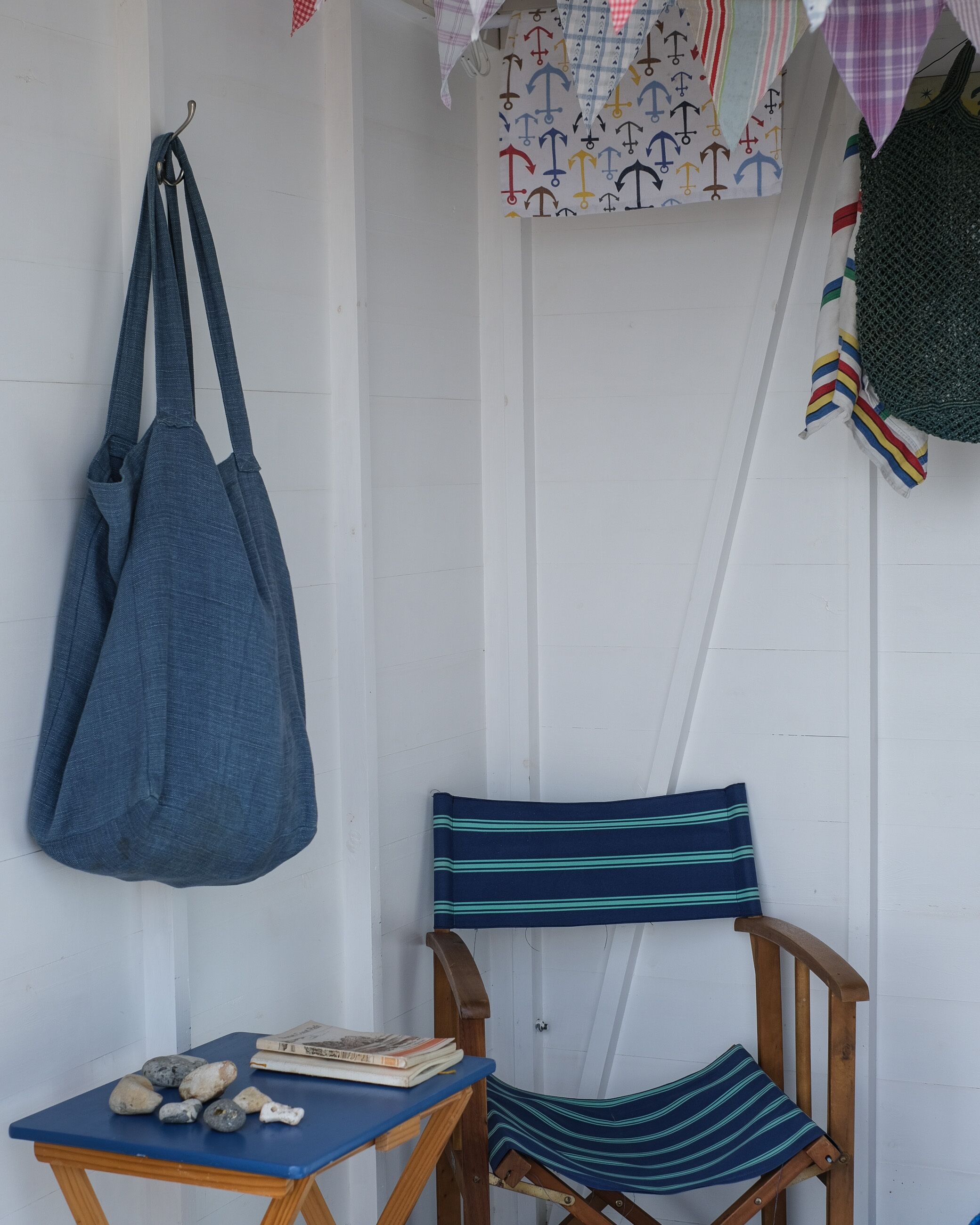 Una silla de madera con un cojín a rayas, una mesita con libros y piedras, y un bolso azul colgado de un gancho.