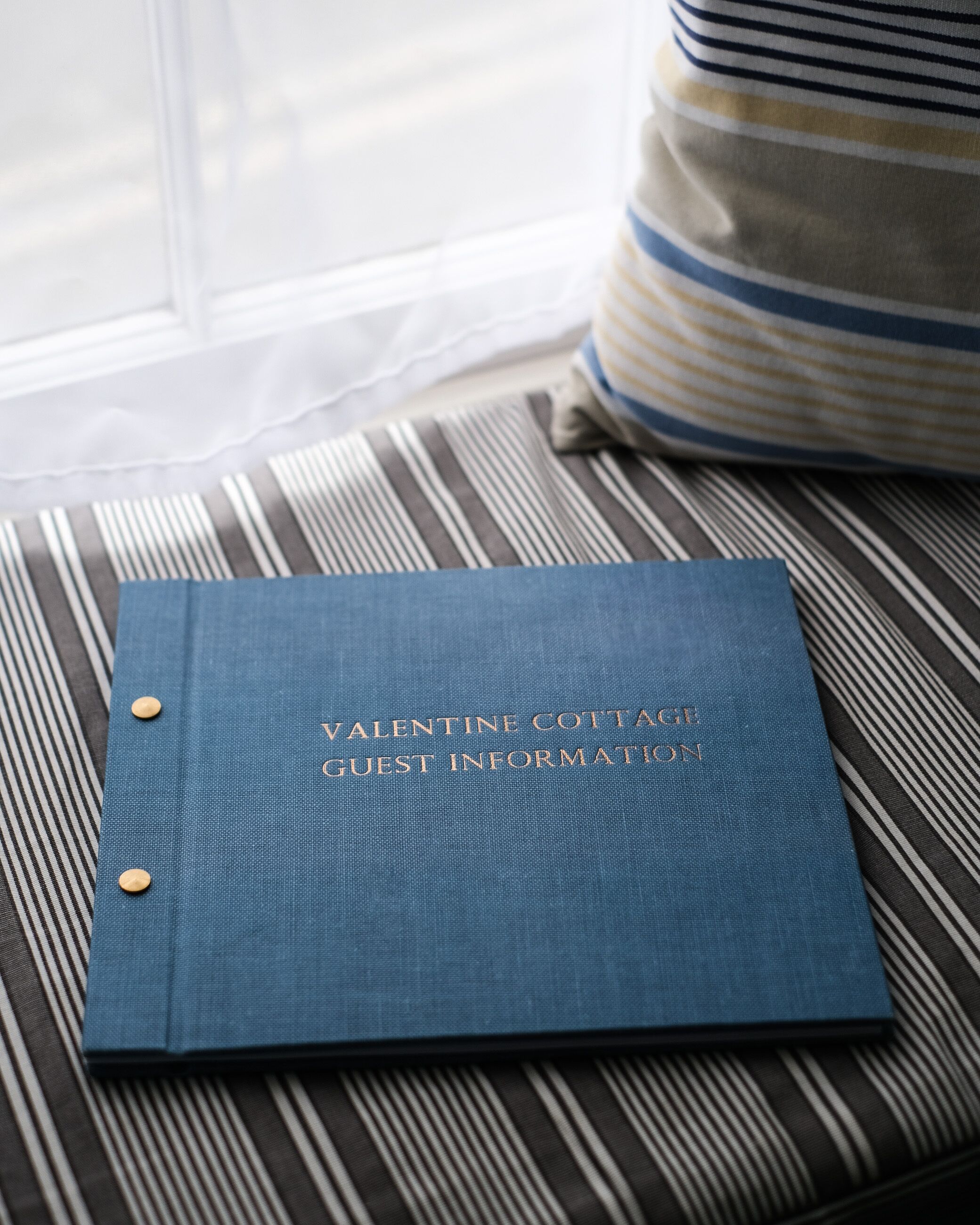 Un classeur bleu portant l’inscription « Informations pour les clients de Valentine Cottage » est posé sur un coussin à rayures.