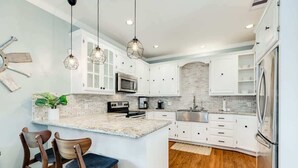A modern kitchen with white cabinets, a granite countertop, and a central island with bar stools. - Sandyland Shores - Screened Porch, Intracoastal & Sea Views! (Surfside Beach)