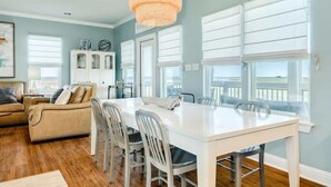 A spacious dining area with a white table and chairs, large windows with blinds, a white cabinet, and a hanging light fixture. - Sandyland Shores - Screened Porch, Intracoastal & Sea Views! (Surfside Beach)