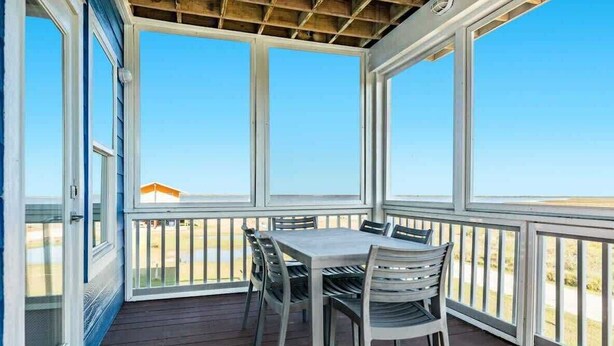 A covered patio with a table and chairs, large windows, and a view of the beach. - Sandyland Shores - Screened Porch, Intracoastal & Sea Views! (Surfside Beach)