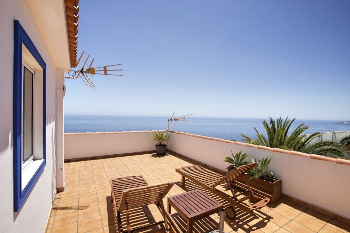 Un balcón con suelo de baldosas, muebles de madera, plantas en macetas y vista al mar.