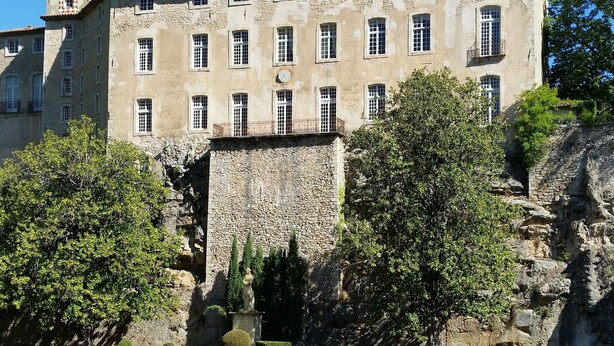 Un edificio histórico con fachada de piedra, múltiples ventanas y un reloj. En frente, un jardín cuidado con setos y un estanque pequeño.