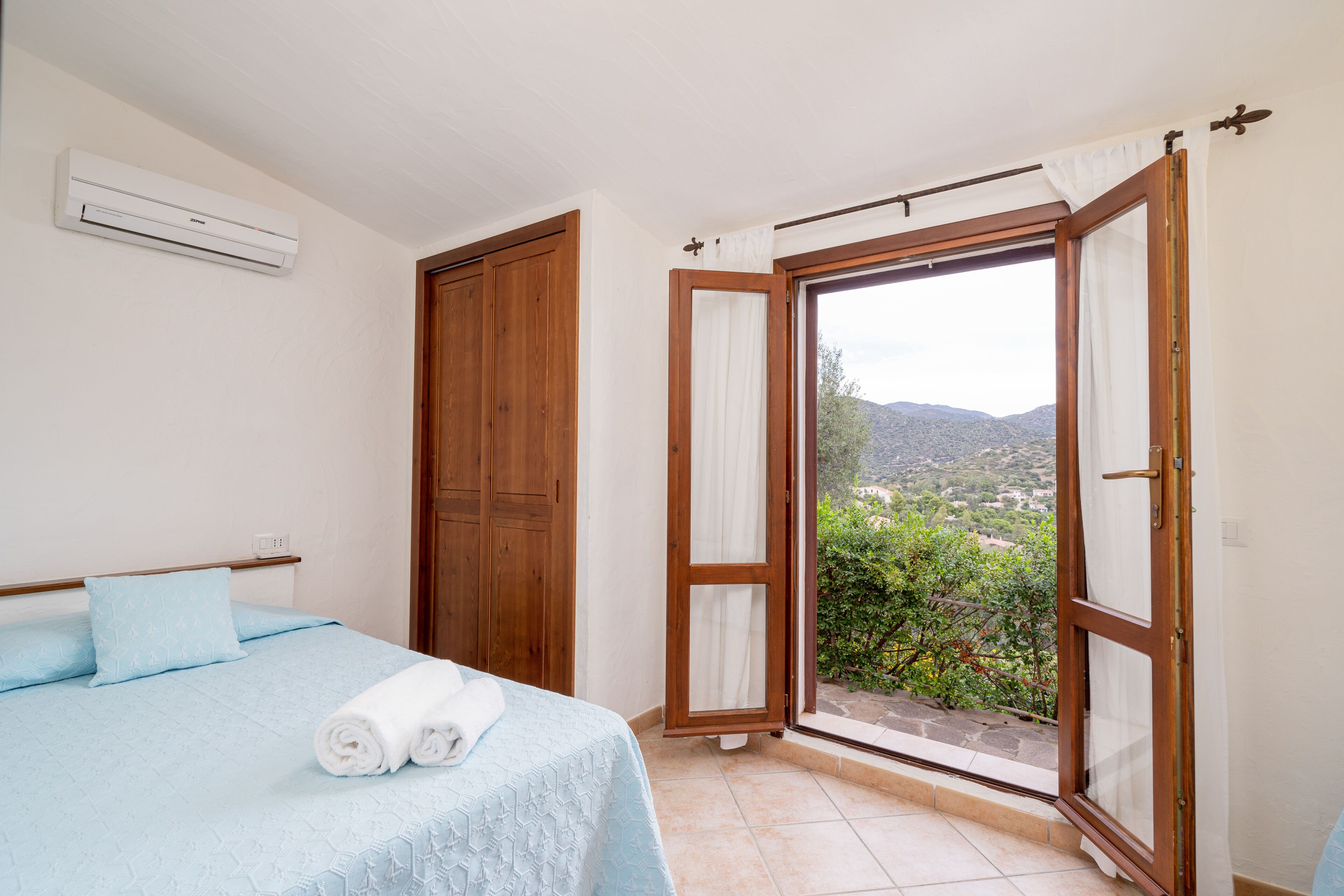 A bedroom with a bed, an air conditioner, a wooden wardrobe, and a sliding glass door leading to a balcony with a view of the landscape.