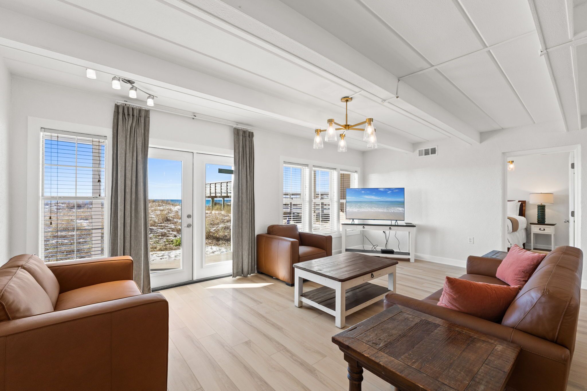 A spacious living room with a large TV, leather sofas, a wooden coffee table, and a balcony with a view of the beach.