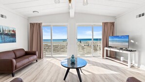 A spacious living room with a large window offering a beach view, a brown leather sofa, a round glass table with a vase, and a TV on a white stand. - CBH A - Where summer is a state of mind! (Fort Walton Beach)